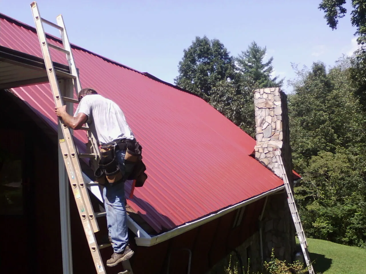 Roofer on a ladder beside a red metal roof and stone chimney — Soffit & Fascia Repair by Roofing Brothers in Laconia